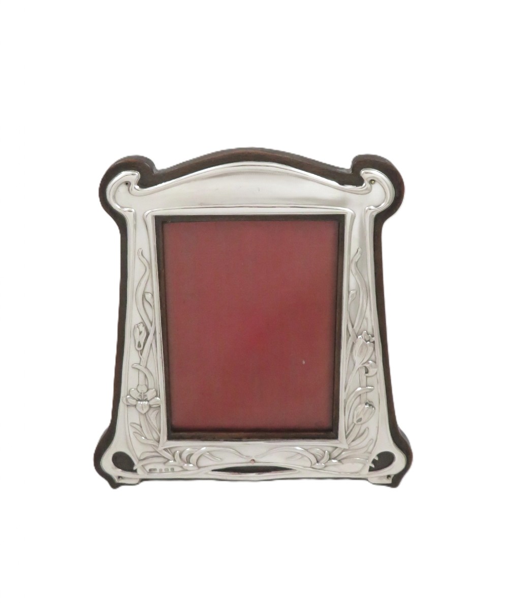 An Edwardian sterling silver Art Nouveau picture frame featuring embossed floral motifs mounted on a dark wood easel backing. View IX