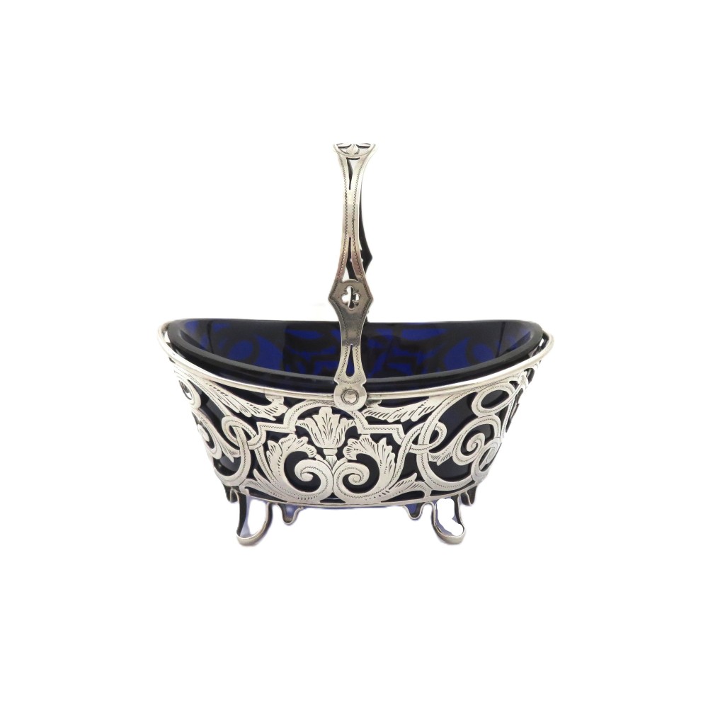 An antique Victorian sterling silver basket with intricate scrollwork piercing, an overhead swing handle, four small feet, and a dark cobalt blue glass liner.