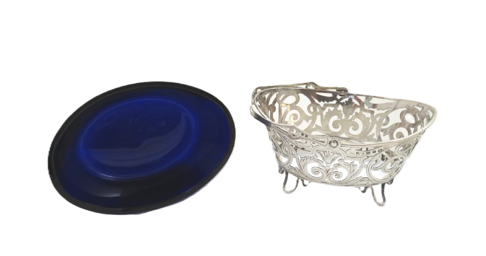 An antique Victorian sterling silver basket with intricate scrollwork piercing, an overhead swing handle, four small feet, and a dark cobalt blue glass liner. View II