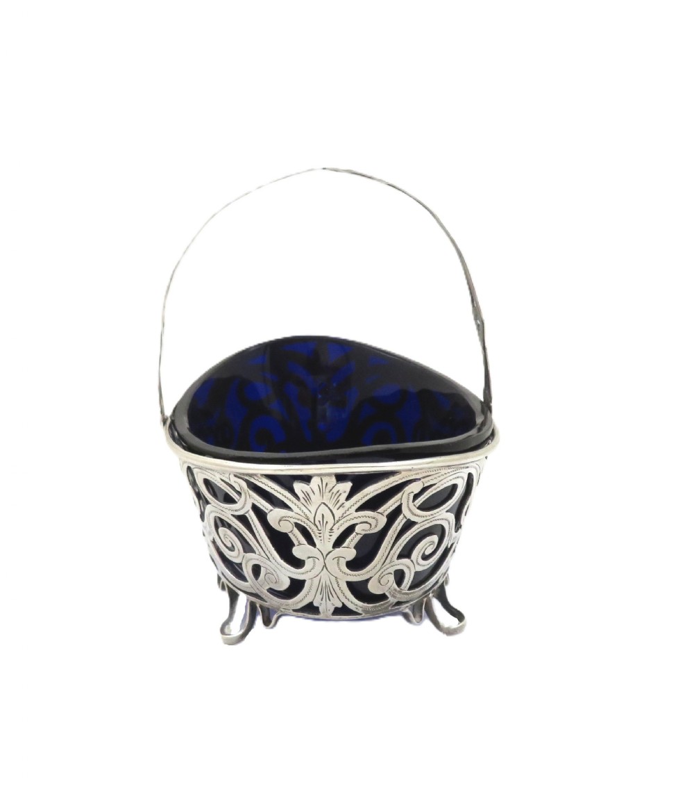 An antique Victorian sterling silver basket with intricate scrollwork piercing, an overhead swing handle, four small feet, and a dark cobalt blue glass liner. View IV