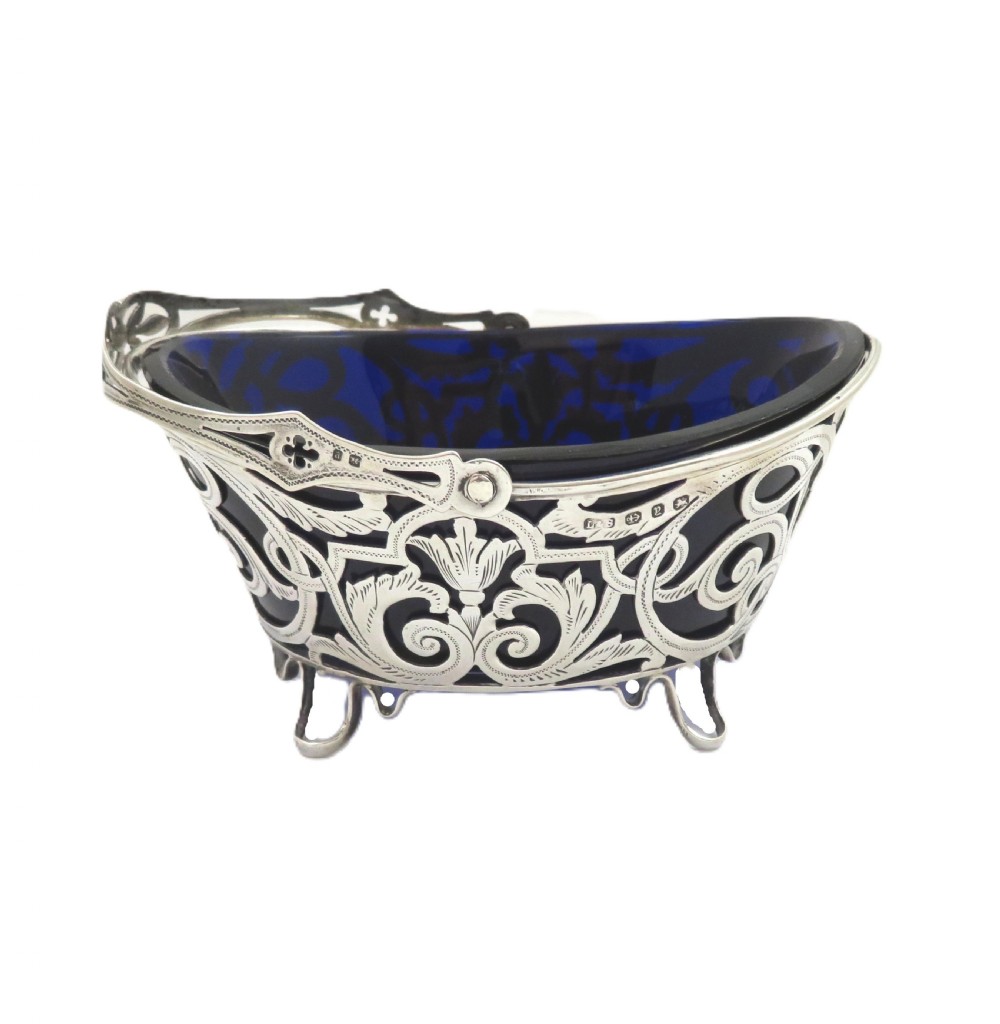 An antique Victorian sterling silver basket with intricate scrollwork piercing, an overhead swing handle, four small feet, and a dark cobalt blue glass liner. View V
