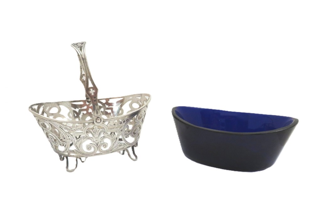 An antique Victorian sterling silver basket with intricate scrollwork piercing, an overhead swing handle, four small feet, and a dark cobalt blue glass liner. View VI