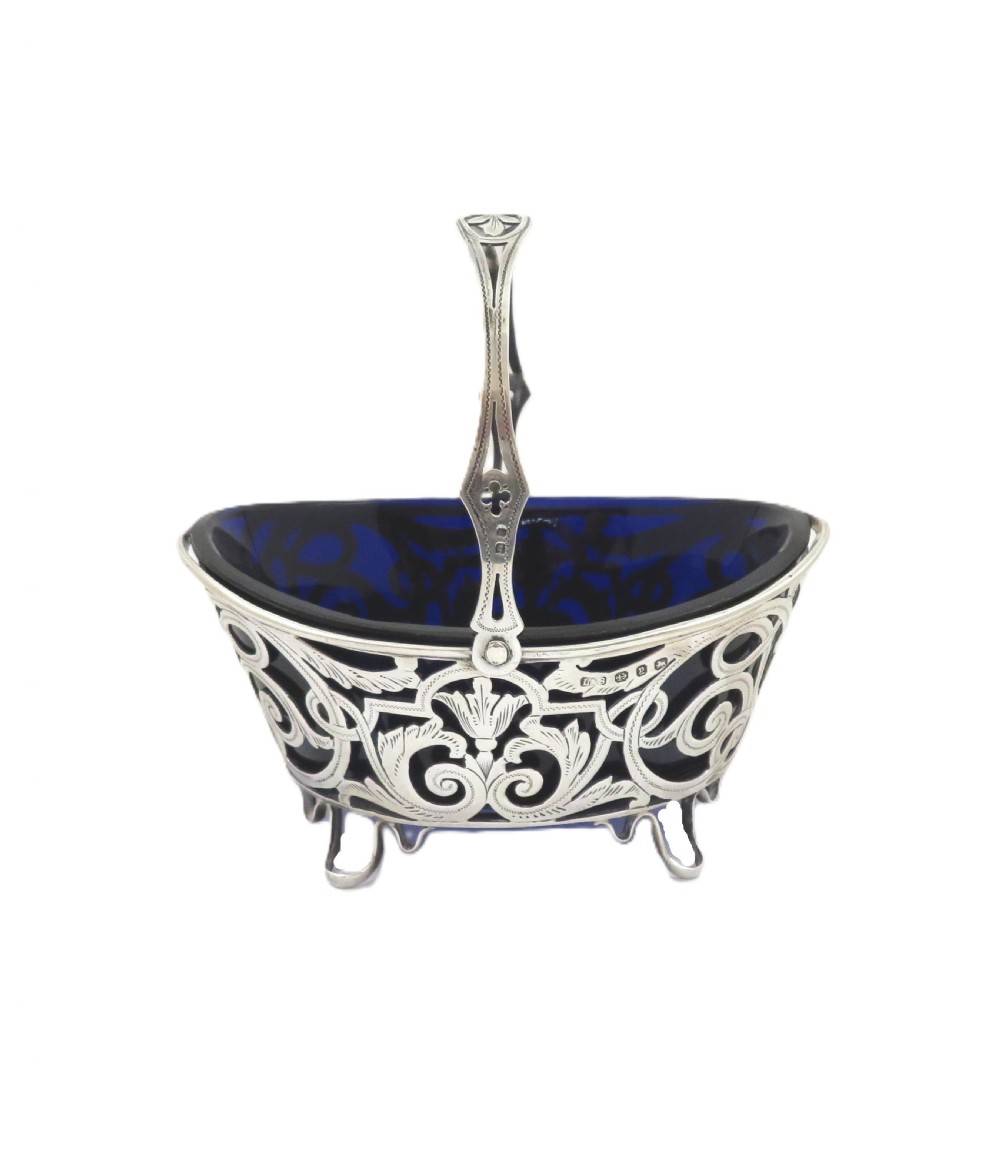 An antique Victorian sterling silver basket with intricate scrollwork piercing, an overhead swing handle, four small feet, and a dark cobalt blue glass liner. View VIII