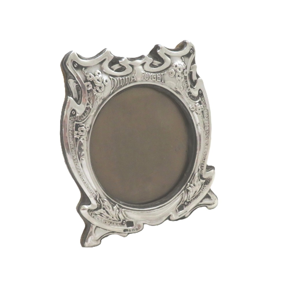 An Art Nouveau sterling silver picture frame featuring embossed floral motifs, sinuous curves, a central circular aperture, and the inscription Dinna Forget.