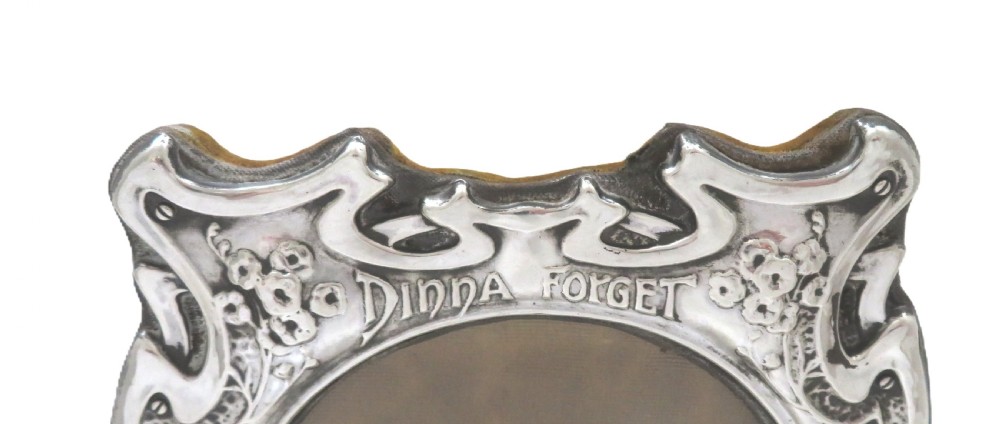 An Art Nouveau sterling silver picture frame featuring embossed floral motifs, sinuous curves, a central circular aperture, and the inscription Dinna Forget. View III