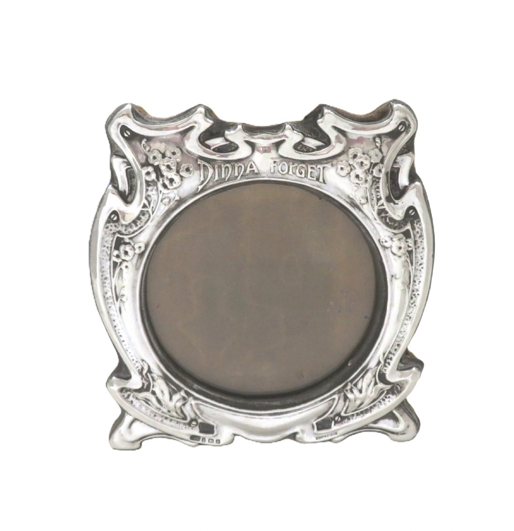 An Art Nouveau sterling silver picture frame featuring embossed floral motifs, sinuous curves, a central circular aperture, and the inscription Dinna Forget. View IV