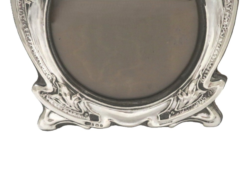 An Art Nouveau sterling silver picture frame featuring embossed floral motifs, sinuous curves, a central circular aperture, and the inscription Dinna Forget. View VI