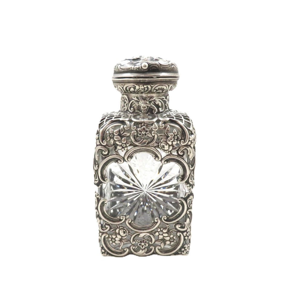 An Edwardian cut glass scent bottle encased in elaborate sterling silver floral scrollwork overlay with a hinged silver cap.