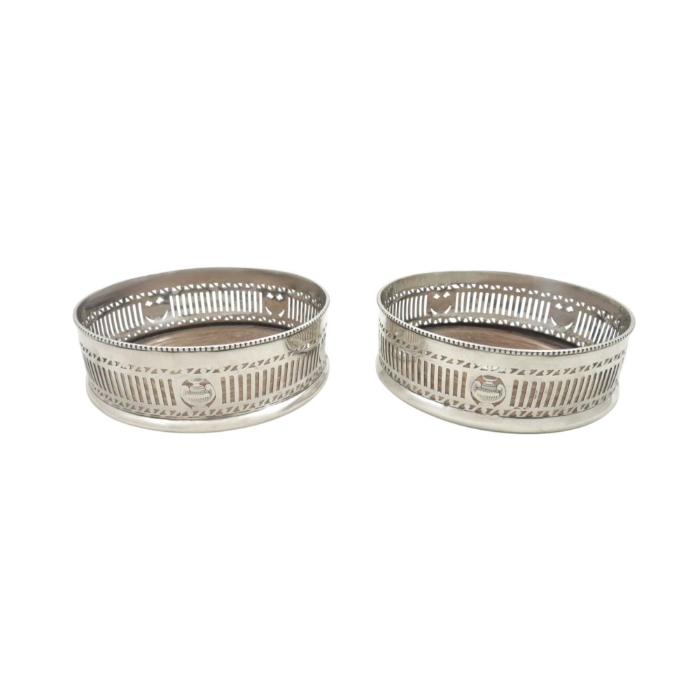 A pair of circular Edwardian sterling silver wine bottle coasters featuring pierced galleries with urn medallions and turned wooden bases.
