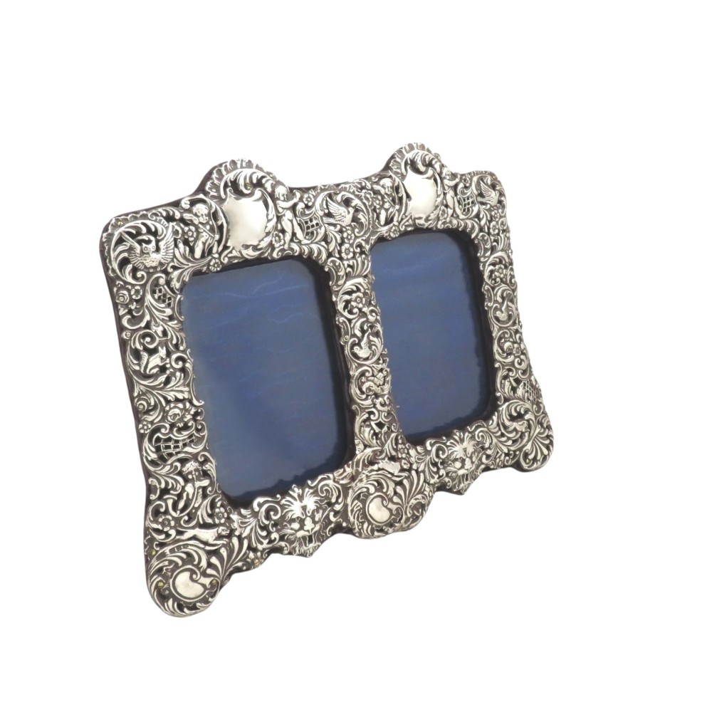 An ornate late Victorian sterling silver double photo frame decorated with embossed scrollwork, foliate motifs, and cherubs.