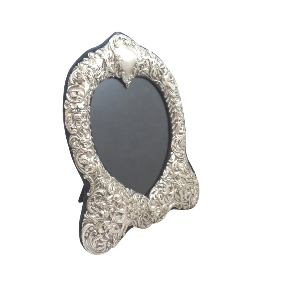 An antique late Victorian sterling silver photo frame featuring a heart-shaped aperture and ornate repousse scrollwork with cherubs.