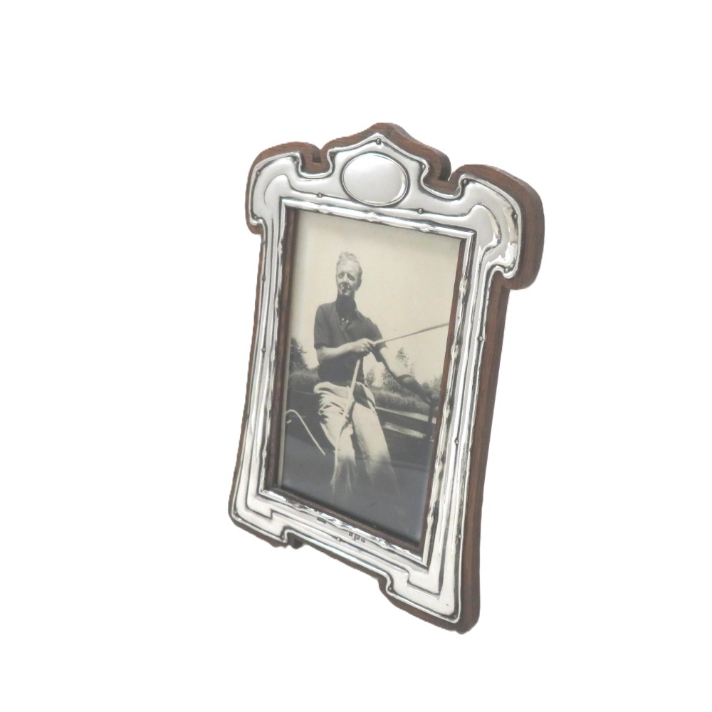 An Art Nouveau sterling silver photo frame with fluid curves mounted on a dark wooden backing.