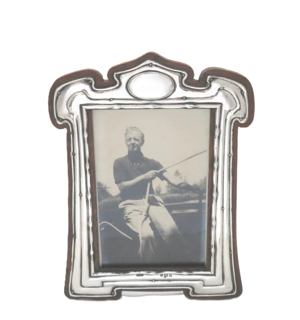 An Art Nouveau sterling silver photo frame with fluid curves mounted on a dark wooden backing. View III