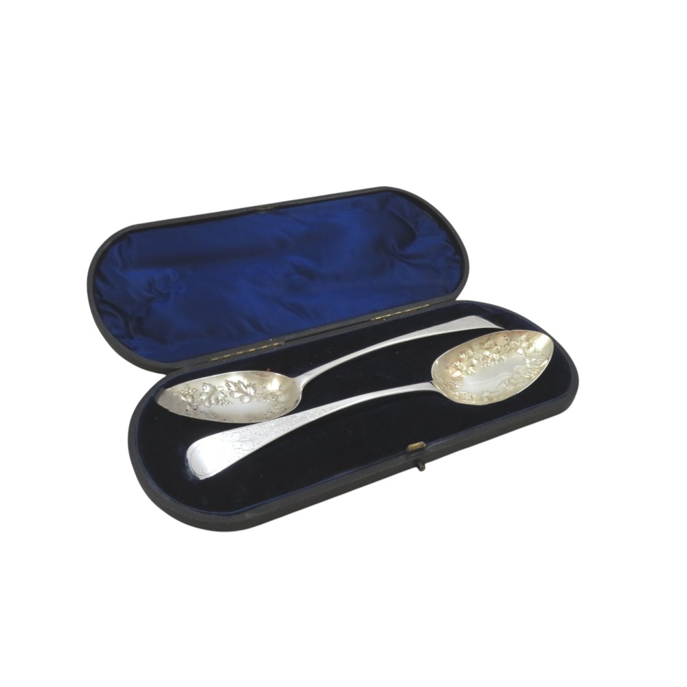 A pair of Georgian sterling silver berry spoons with repousse fruit motifs in their bowls, displayed inside an open blue velvet-lined presentation case.