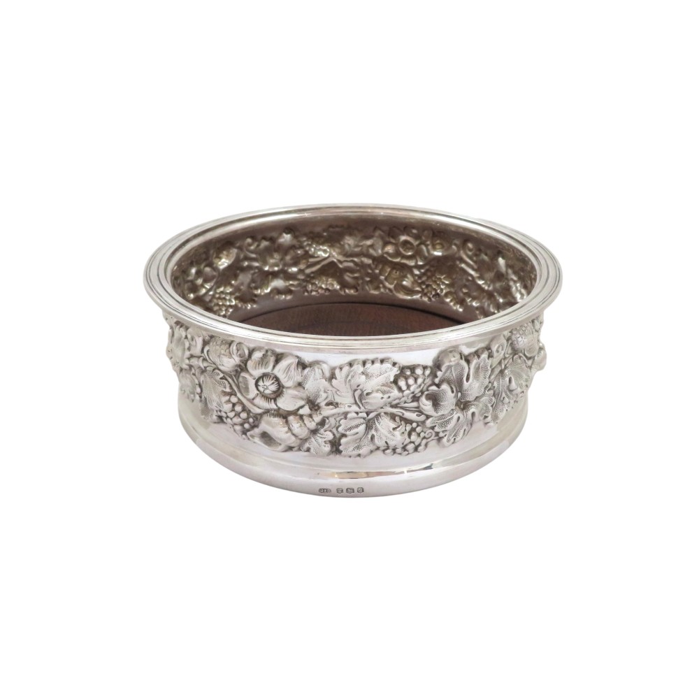 A circular sterling silver wine coaster featuring an embossed grape and vine relief border around a turned wooden base.