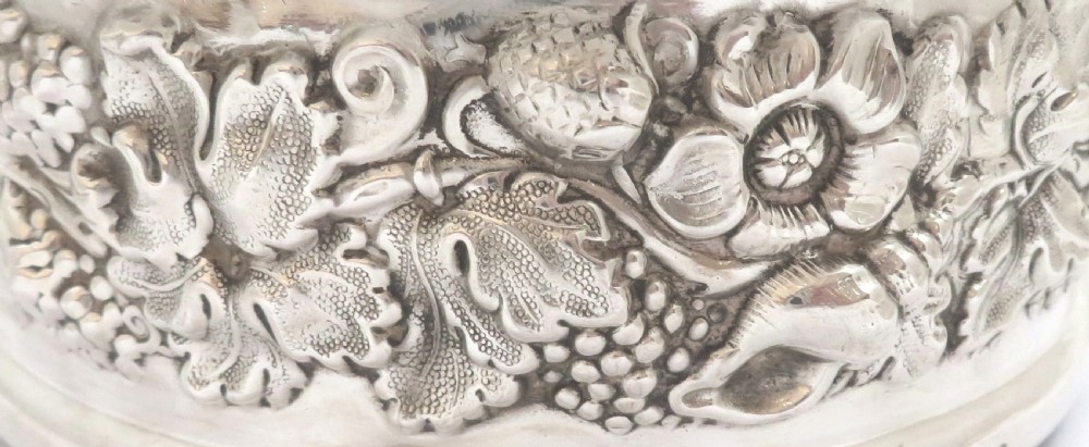 A circular sterling silver wine coaster featuring an embossed grape and vine relief border around a turned wooden base. View V