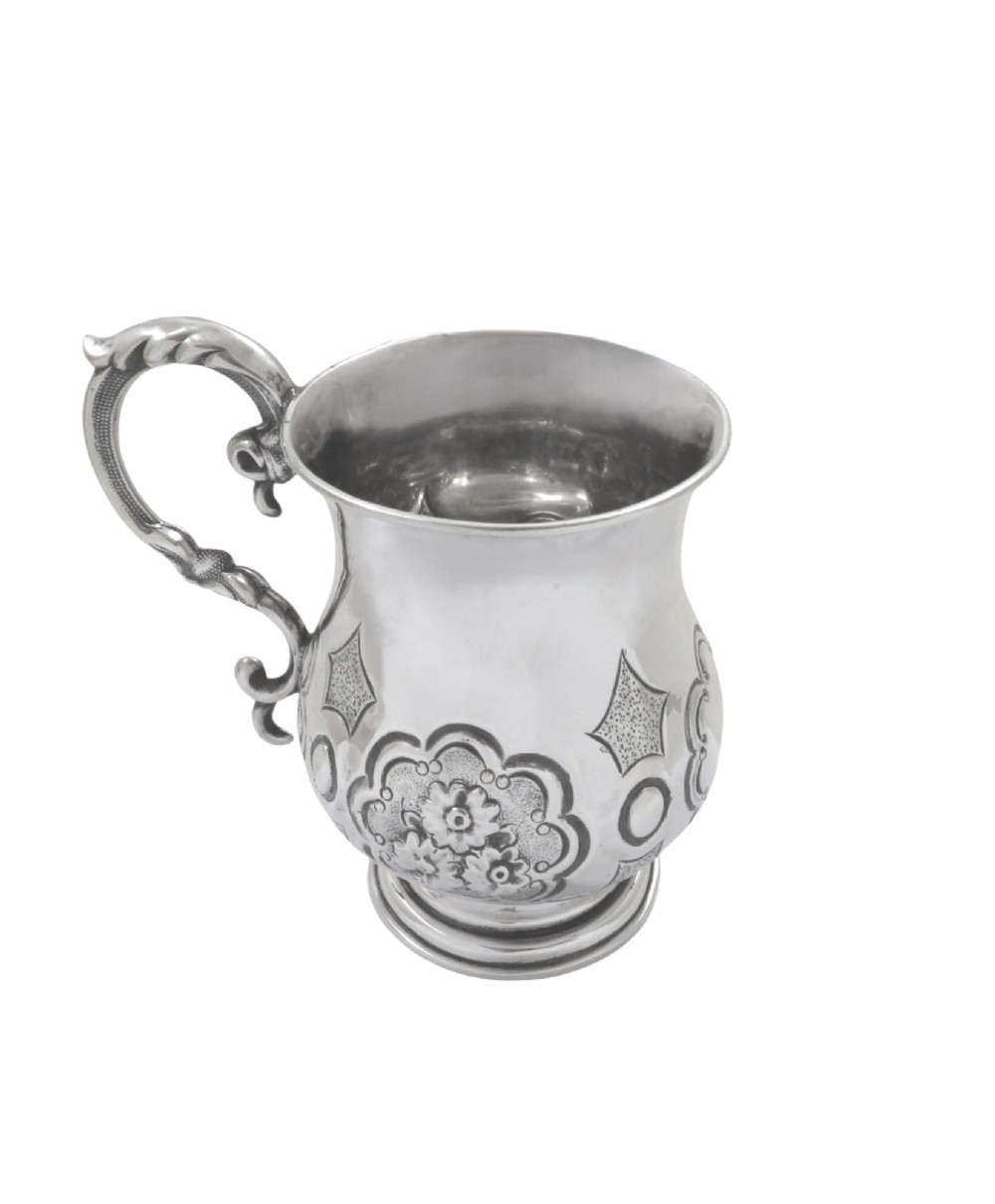 A Victorian sterling silver christening mug featuring floral chasing, geometric embossing, and a scroll handle. View V