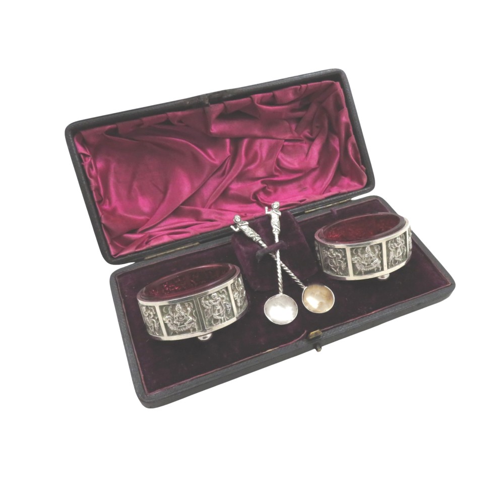 A pair of circular sterling silver salt cellars with red glass liners and matching spoons featuring Hindu deity relief panels, housed inside a velvet-lined presentation case.