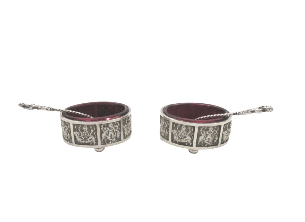 A pair of circular sterling silver salt cellars with red glass liners and matching spoons featuring Hindu deity relief panels, housed inside a velvet-lined presentation case. View III
