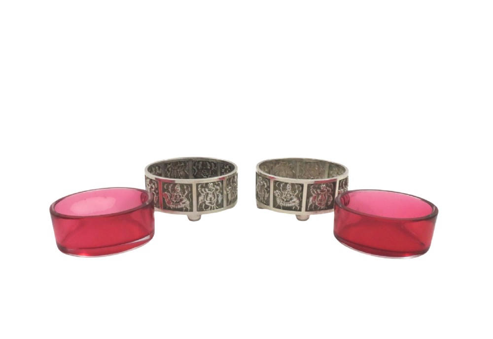 A pair of circular sterling silver salt cellars with red glass liners and matching spoons featuring Hindu deity relief panels, housed inside a velvet-lined presentation case. View VII