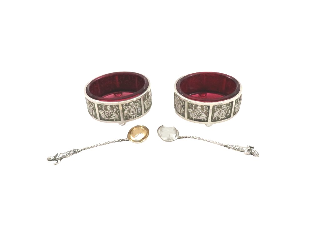 A pair of circular sterling silver salt cellars with red glass liners and matching spoons featuring Hindu deity relief panels, housed inside a velvet-lined presentation case. View IX