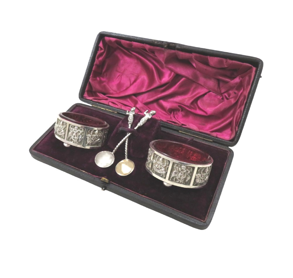 A pair of circular sterling silver salt cellars with red glass liners and matching spoons featuring Hindu deity relief panels, housed inside a velvet-lined presentation case. View X