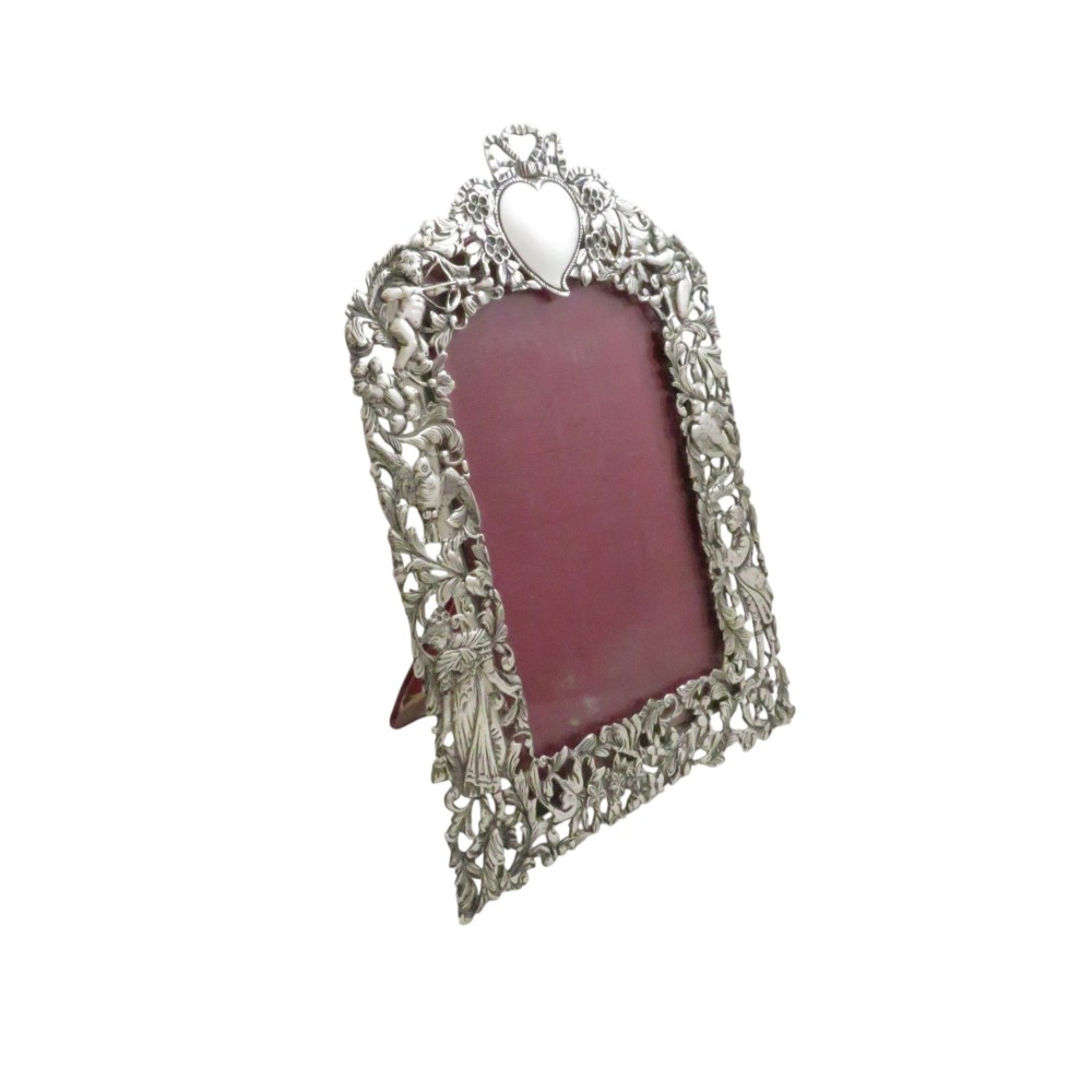 An ornate late Victorian sterling silver easel-back photo frame decorated with pierced cherubs, birds, and floral scrollwork, fitted with a burgundy velvet backing.