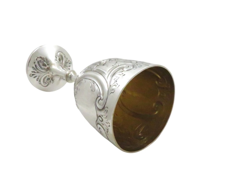 An Edwardian sterling silver wine goblet featuring scroll repousse engraving, an unengraved central cartouche, a knopped stem, and a gilt interior. View V