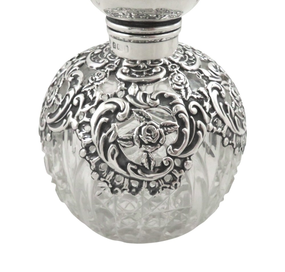 An Edwardian clear glass scent bottle featuring a round body with ornate embossed sterling silver overlay and matching cap. View II