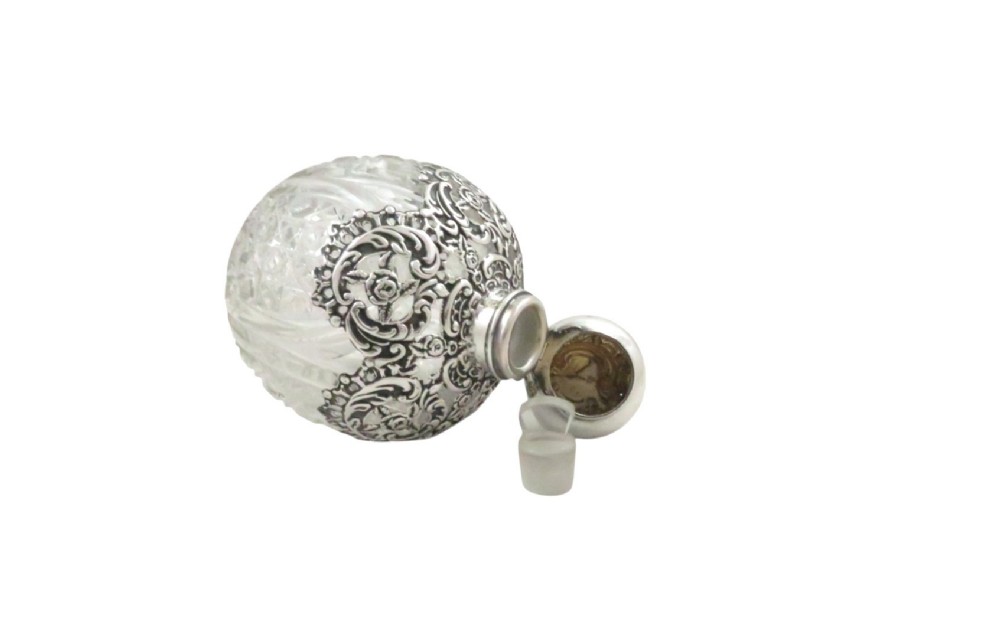 An Edwardian clear glass scent bottle featuring a round body with ornate embossed sterling silver overlay and matching cap. View III