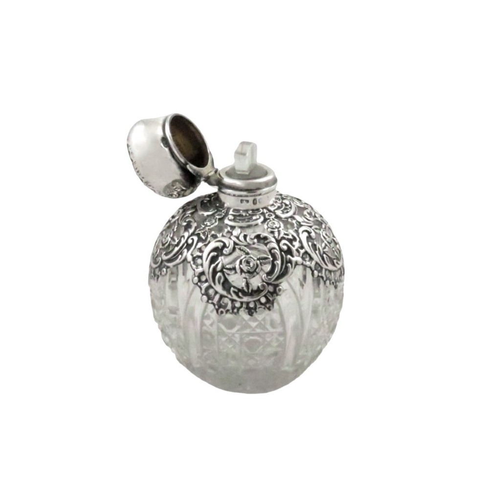 An Edwardian clear glass scent bottle featuring a round body with ornate embossed sterling silver overlay and matching cap. View V