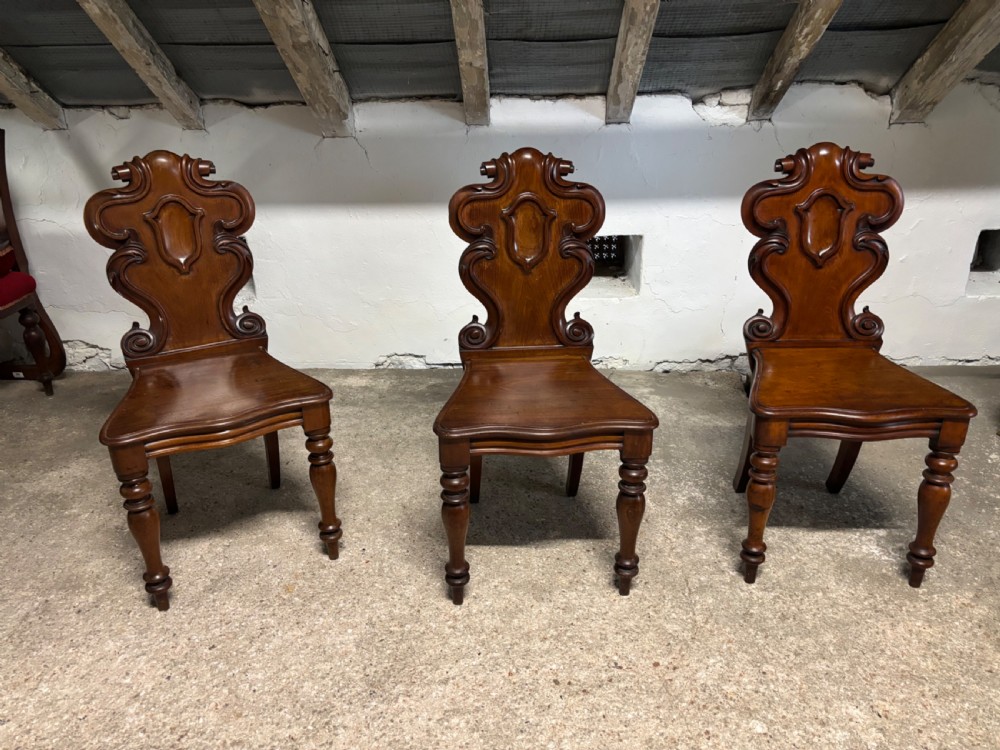 A set of three Victorian mahogany hall chairs with carved scrollwork backs, central shield motifs, solid timber seats, and turned front legs.