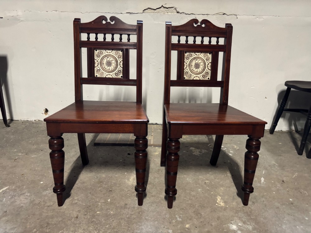 A pair of Victorian mahogany hall chairs featuring carved scroll crests, spindled upper rails, decorative ceramic tile back inserts, solid timber seats, and turned front legs.