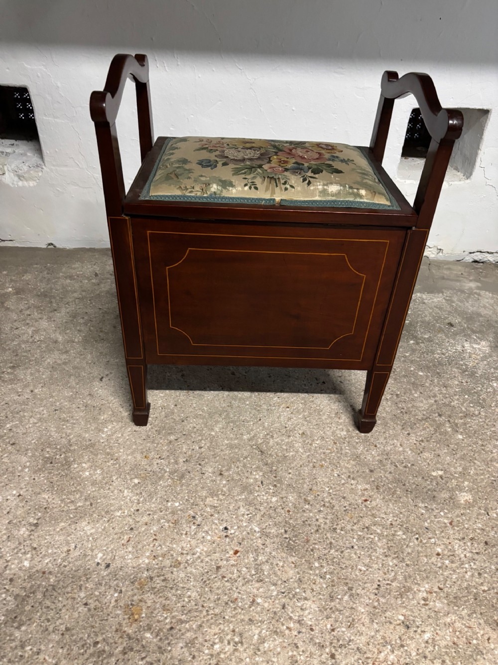 An Edwardian mahogany piano stool featuring linear boxwood inlay, twin raised side handles, and a hinged floral tapestry seat.