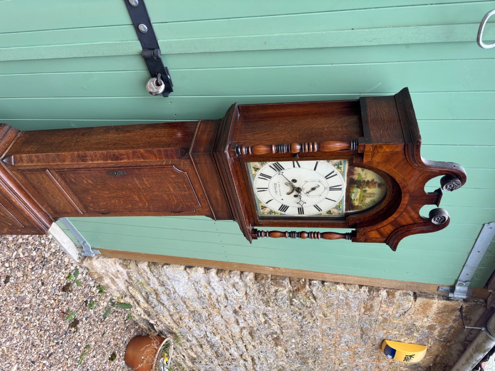 An early 19th-century oak longcase clock featuring a swan-neck pediment, a painted dial with Roman numerals, and a panelled trunk. View V