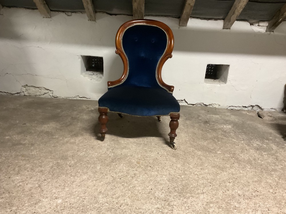 A Victorian low-slung nursing chair featuring a carved mahogany frame, navy blue velvet upholstery, a spoon-shaped backrest, and turned front legs on castors.