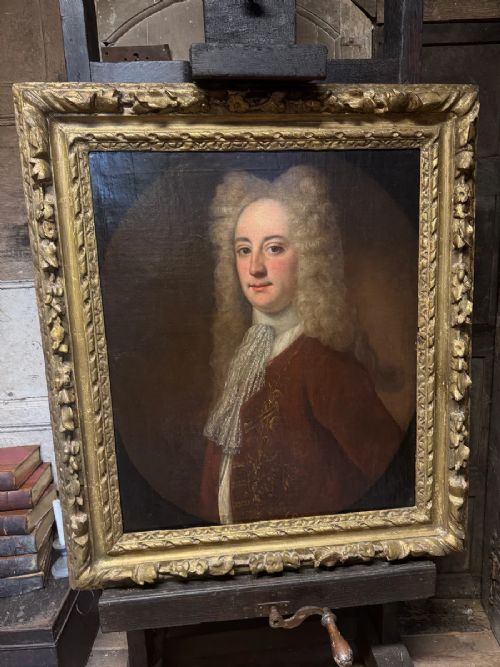 An early 18th-century oil portrait depicting a gentleman in a crimson coat and powdered wig, set within a heavily carved gilded frame. View VII