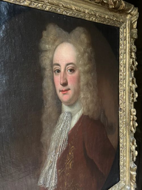 An early 18th-century oil portrait depicting a gentleman in a crimson coat and powdered wig, set within a heavily carved gilded frame. View VIII