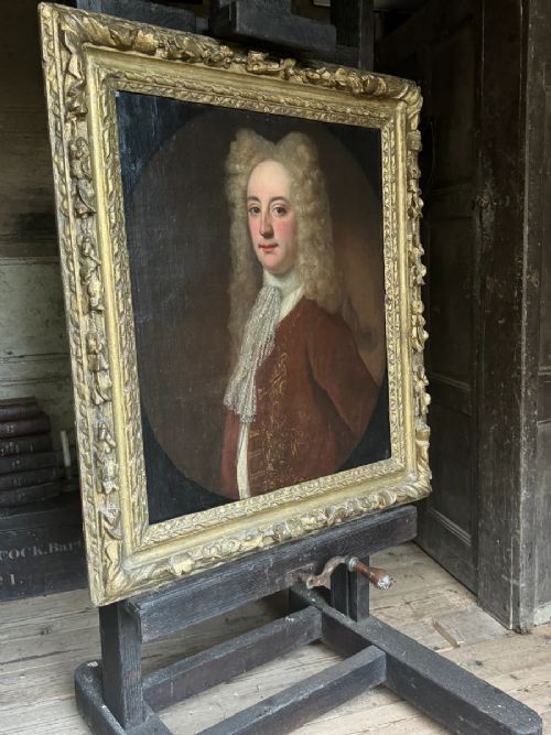 An early 18th-century oil portrait depicting a gentleman in a crimson coat and powdered wig, set within a heavily carved gilded frame. View IX
