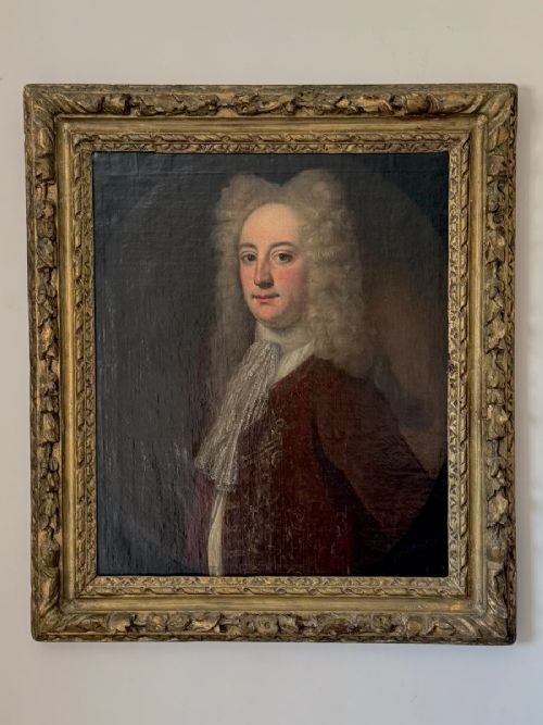An early 18th-century oil portrait depicting a gentleman in a crimson coat and powdered wig, set within a heavily carved gilded frame. View X