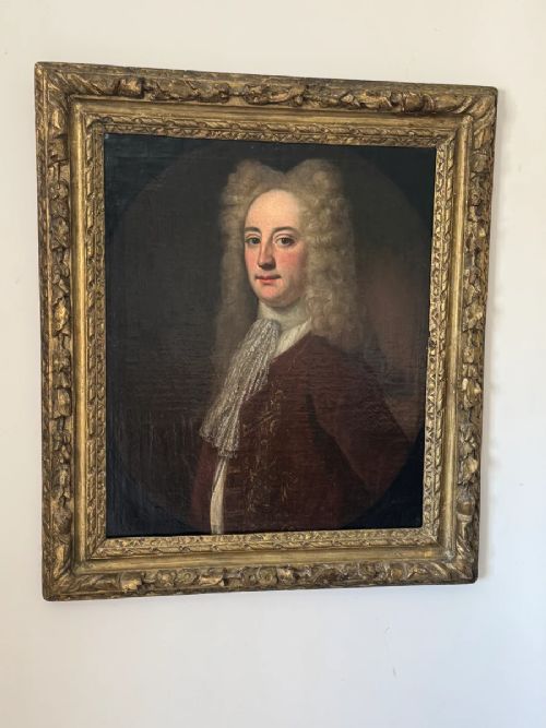 An early 18th-century oil portrait depicting a gentleman in a crimson coat and powdered wig, set within a heavily carved gilded frame. View XII