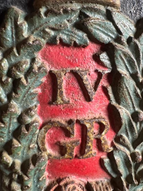 fig 2. A vertical rectangular cast iron plaque featuring a painted crown, laurel wreath surrounding the letters IV GR, and the initials B O at the base.
