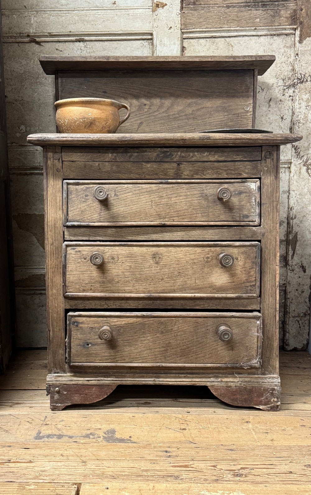 A small early 19th-century primitive oak chest featuring three drawers with turned wooden knobs and a raised wooden back shelf. View V