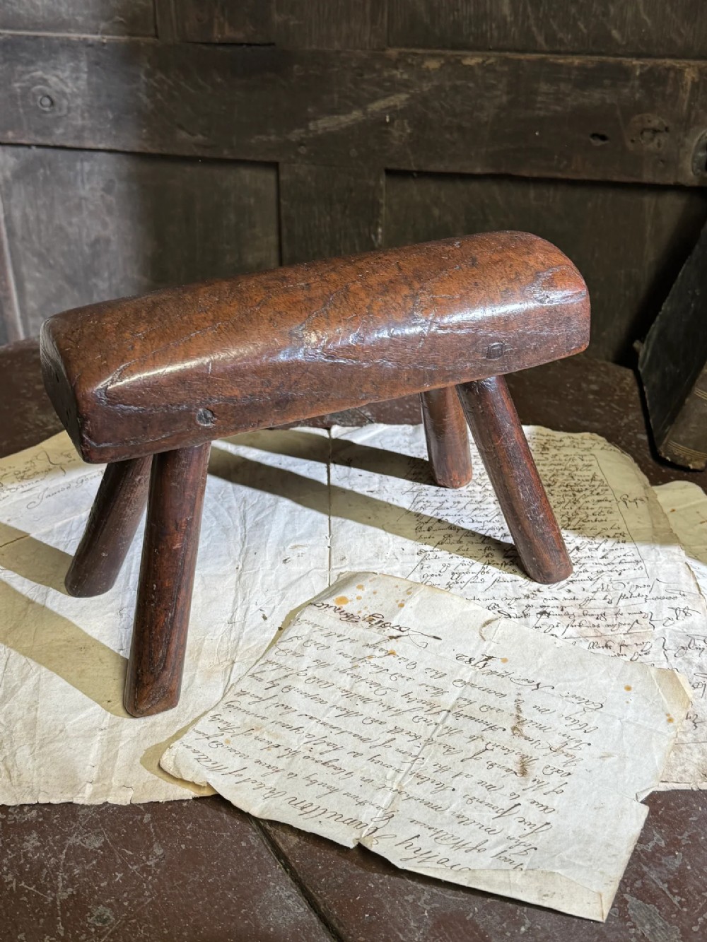 A small antique dark timber vernacular stool with a sculpted rounded top and four splayed legs.