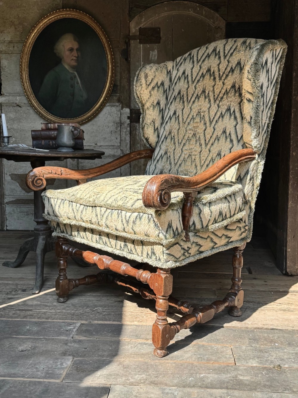 An early eighteenth-century French oak wingback armchair featuring patterned fabric upholstery, carved scroll armrests, and turned legs with stretchers.
