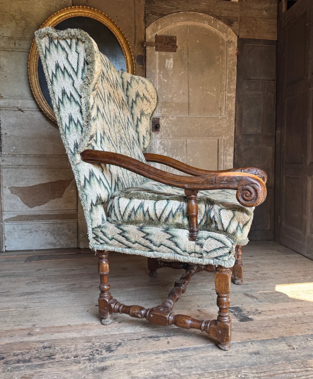 An early eighteenth-century French oak wingback armchair featuring patterned fabric upholstery, carved scroll armrests, and turned legs with stretchers. View II