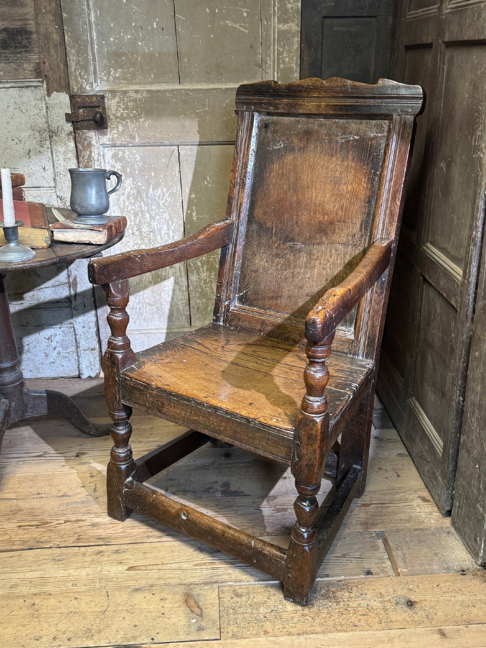 A 17th-century solid dark oak wainscot armchair featuring a panelled backrest, turned front legs, and wooden armrests.