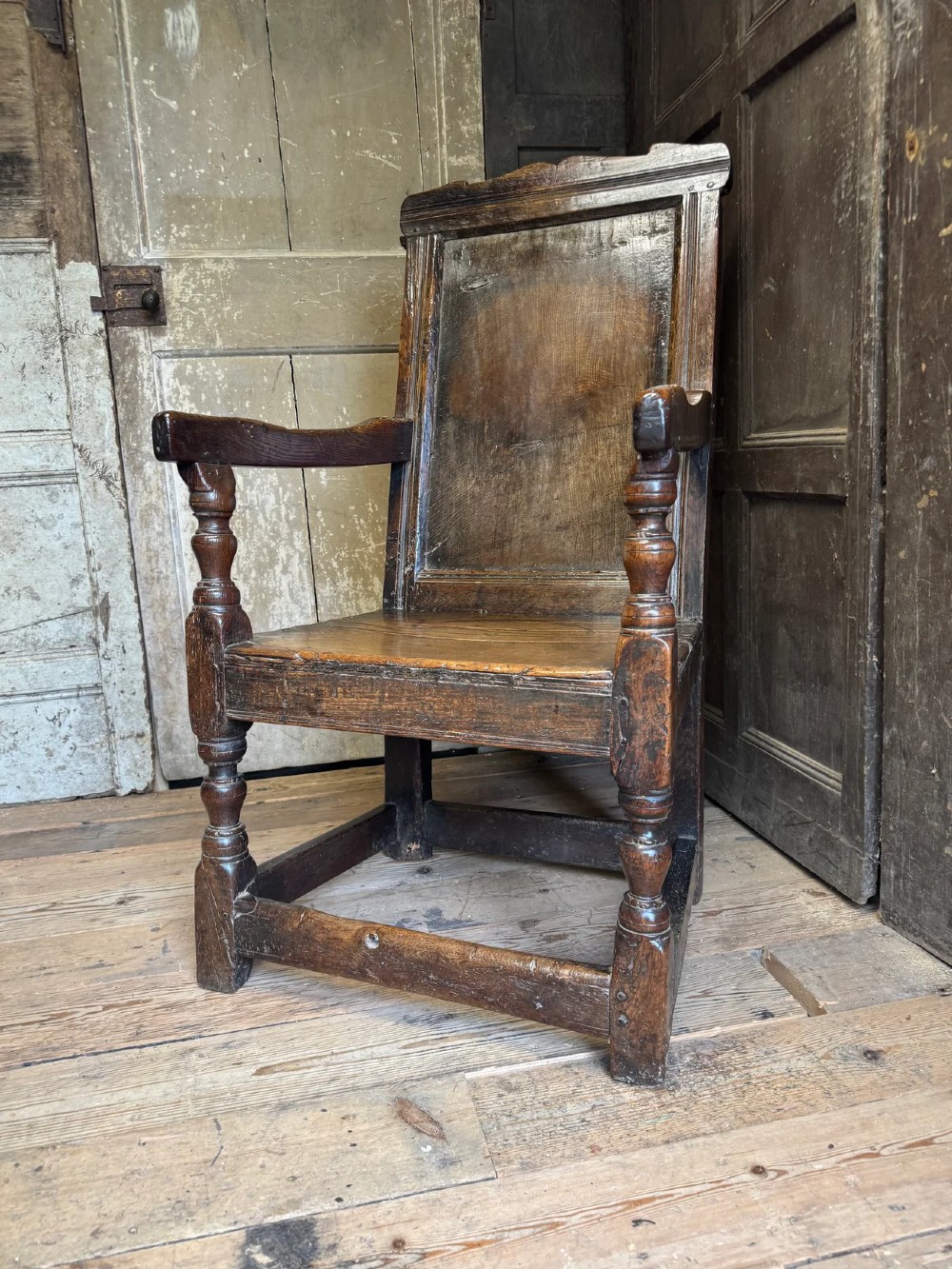 A 17th-century solid dark oak wainscot armchair featuring a panelled backrest, turned front legs, and wooden armrests. View IX