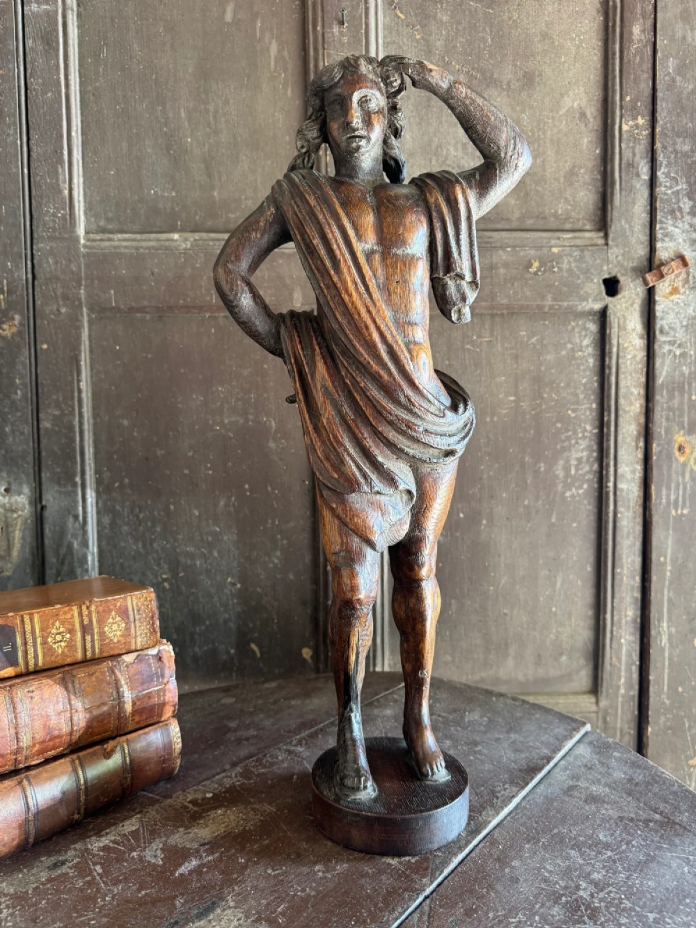 An 18th-century carved oak standing male figure with drapery across his shoulder set on a rounded base.