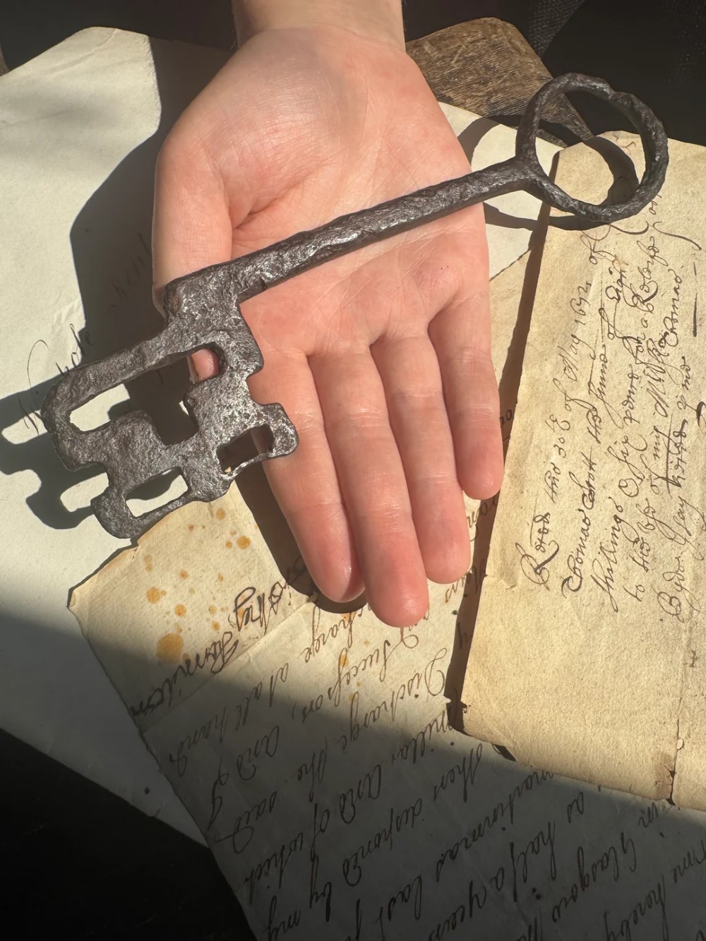 A hand-forged dark wrought iron key featuring a circular bow, a straight shaft, and a cut bit resting on manuscript paper. View III